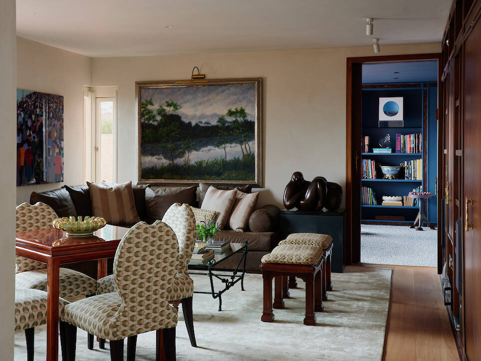 Living room with a brown leather sectional, a dining table with patterned chairs, and a landscape painting above the sofa, leading to a blue room with bookshelves
