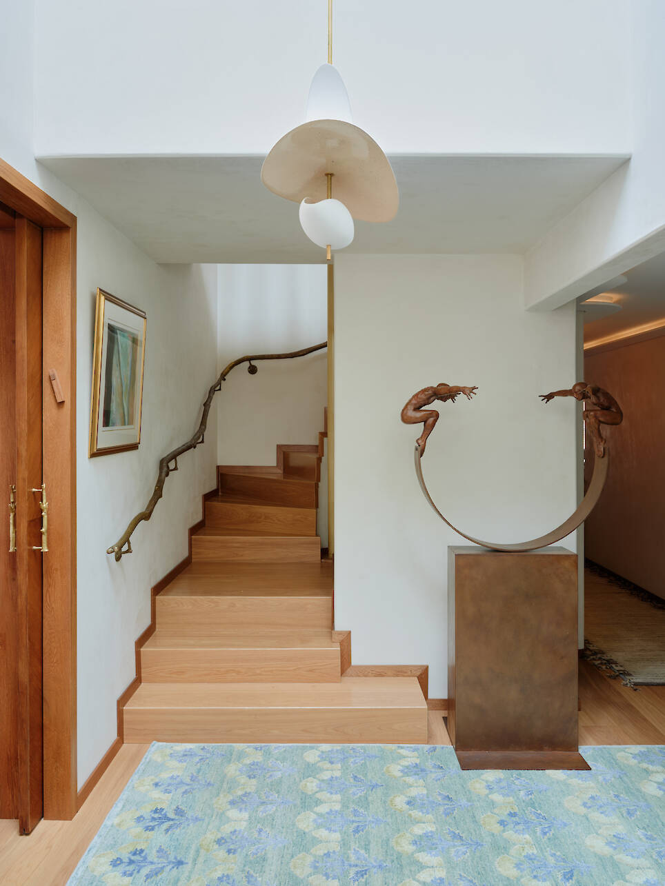 A wooden staircase with a branch-like handrail, featuring a sculpture of two figures on a curved metal base in the foreground