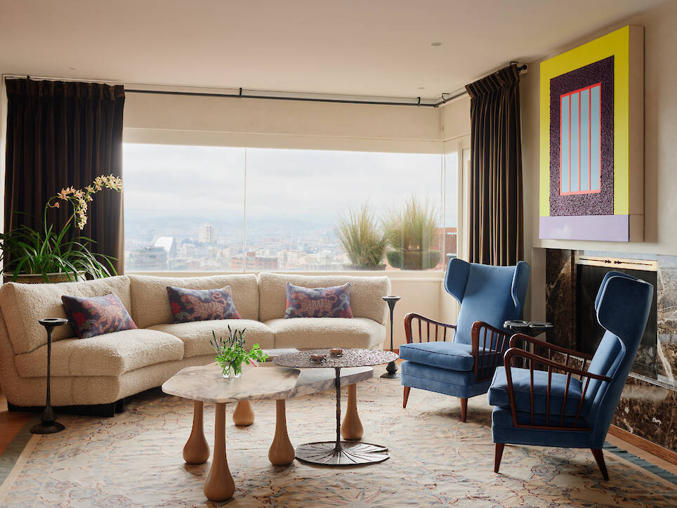 A living room with a curved, cream-colored sofa, two blue armchairs, and two tiered coffee tables in front of a window overlooking a city