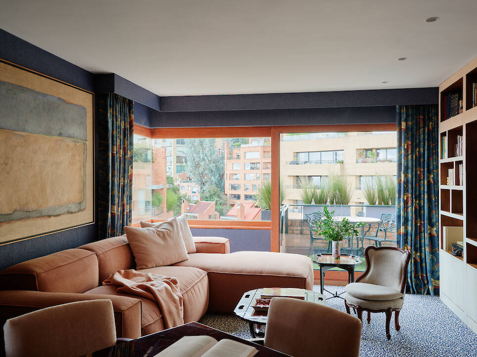 A living room with an orange sectional sofa, a patterned armchair, a built-in bookshelf, and a large window overlooking a balcony and city buildings