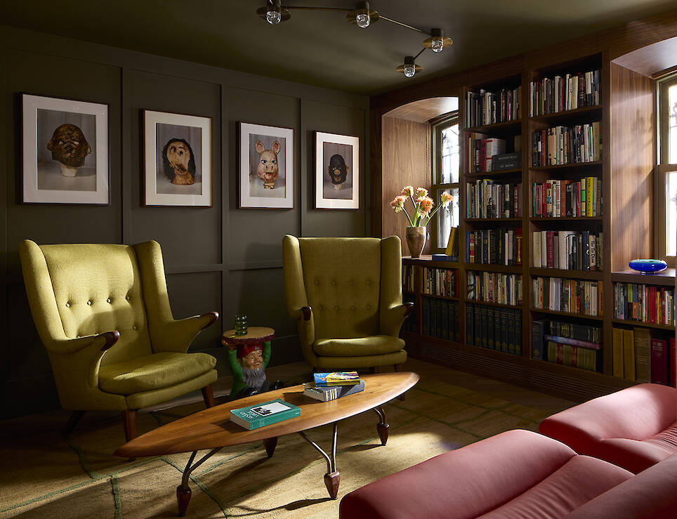 Two lime green wingback chairs face a wooden coffee table in a library with floor-to-ceiling bookshelves and four framed portrait prints on the wall