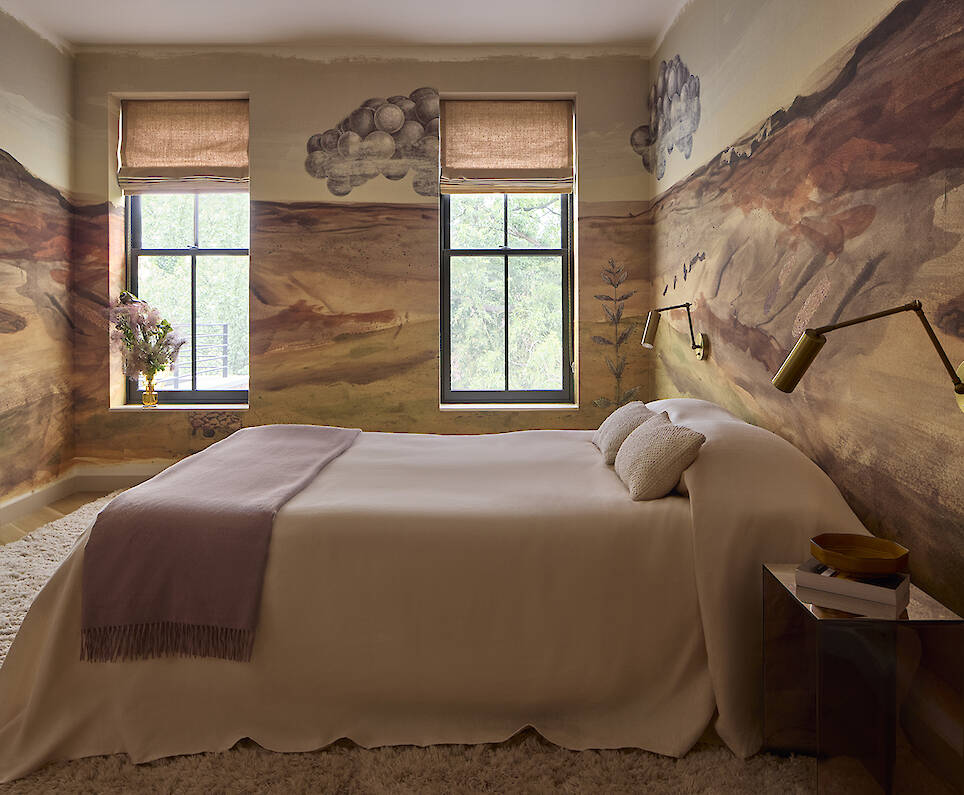 A bedroom with walls featuring a landscape mural, a bed with a light-colored cover and a mauve throw, two windows with tan shades, and a mirrored nightstand