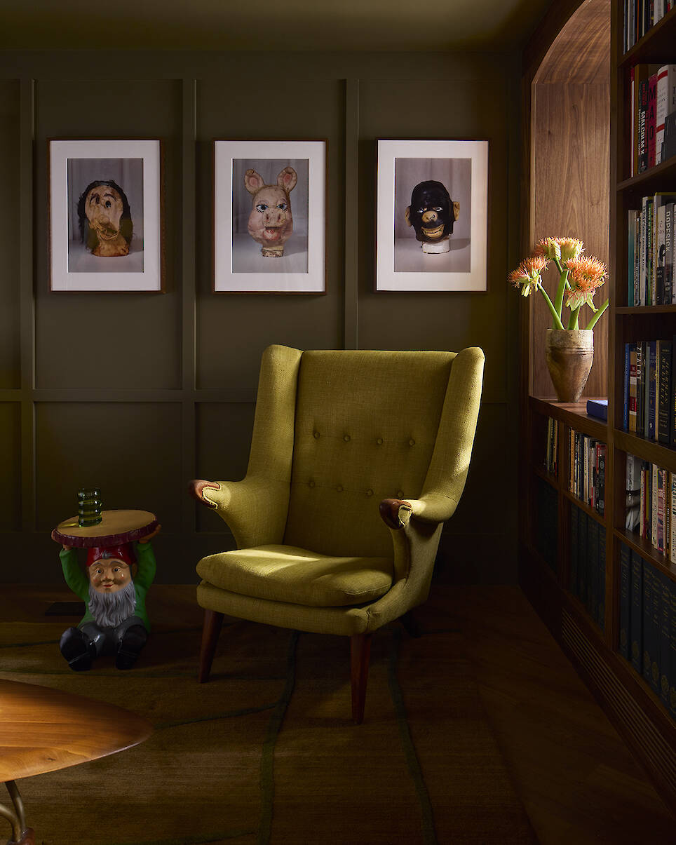 A yellow-green wingback chair sits in a room with three framed masks on the wall, a gnome side table, and a built-in bookshelf