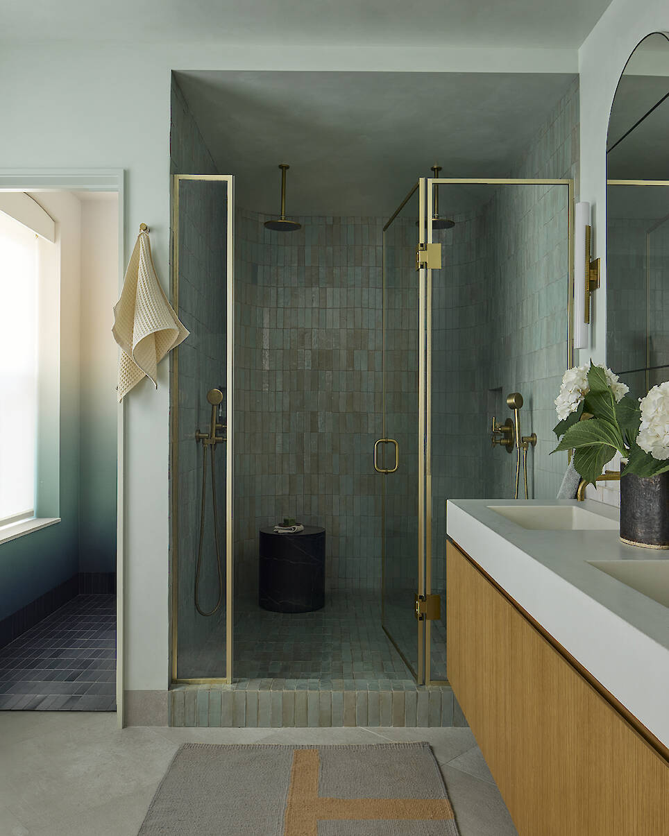 A walk-in shower with green tiled walls, a glass door, and gold fixtures, next to a double vanity with a wooden cabinet