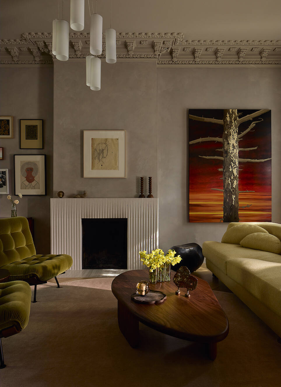Living room with a fireplace, a large abstract painting of a tree, olive green armchairs, and a curved wooden coffee table with a vase of yellow flowers