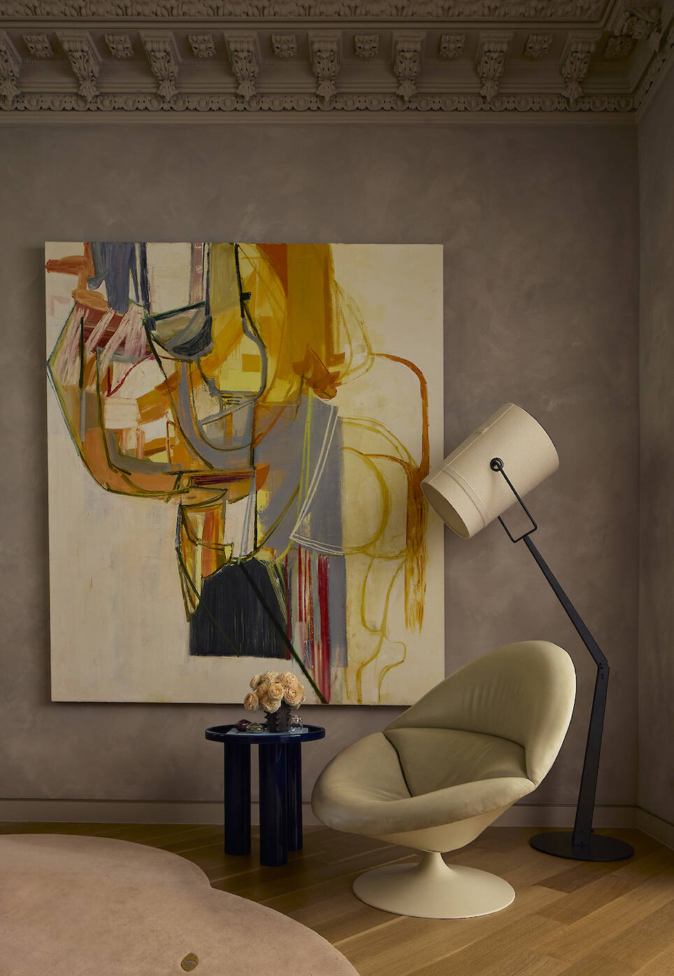 A large abstract painting with orange and yellow brushstrokes hangs behind a cream-colored swivel chair, a blue side table with a flower bouquet, and a floor lamp