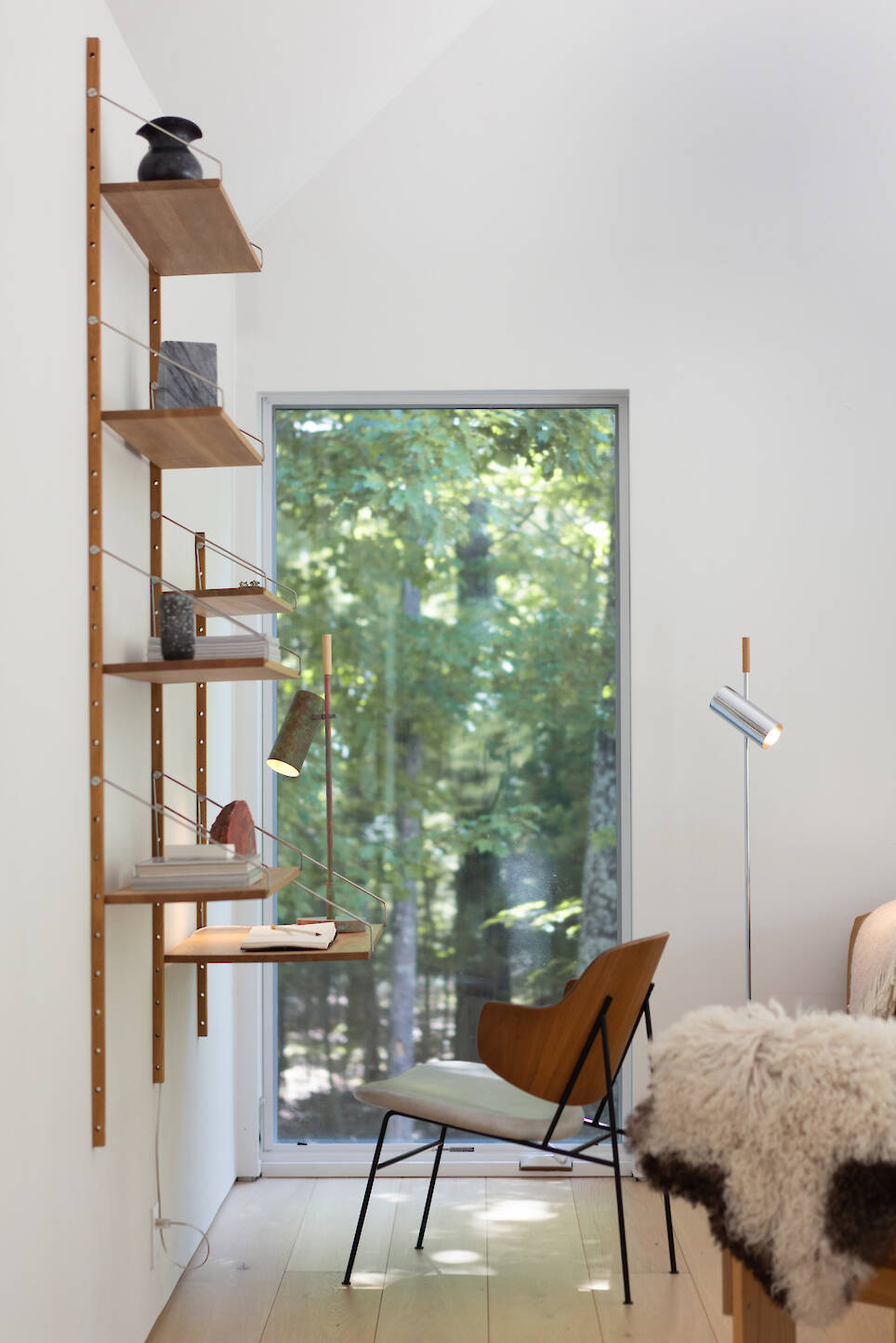 A modular wall-mounted shelving unit with a desk surface sits beside a vertical window and a wooden chair