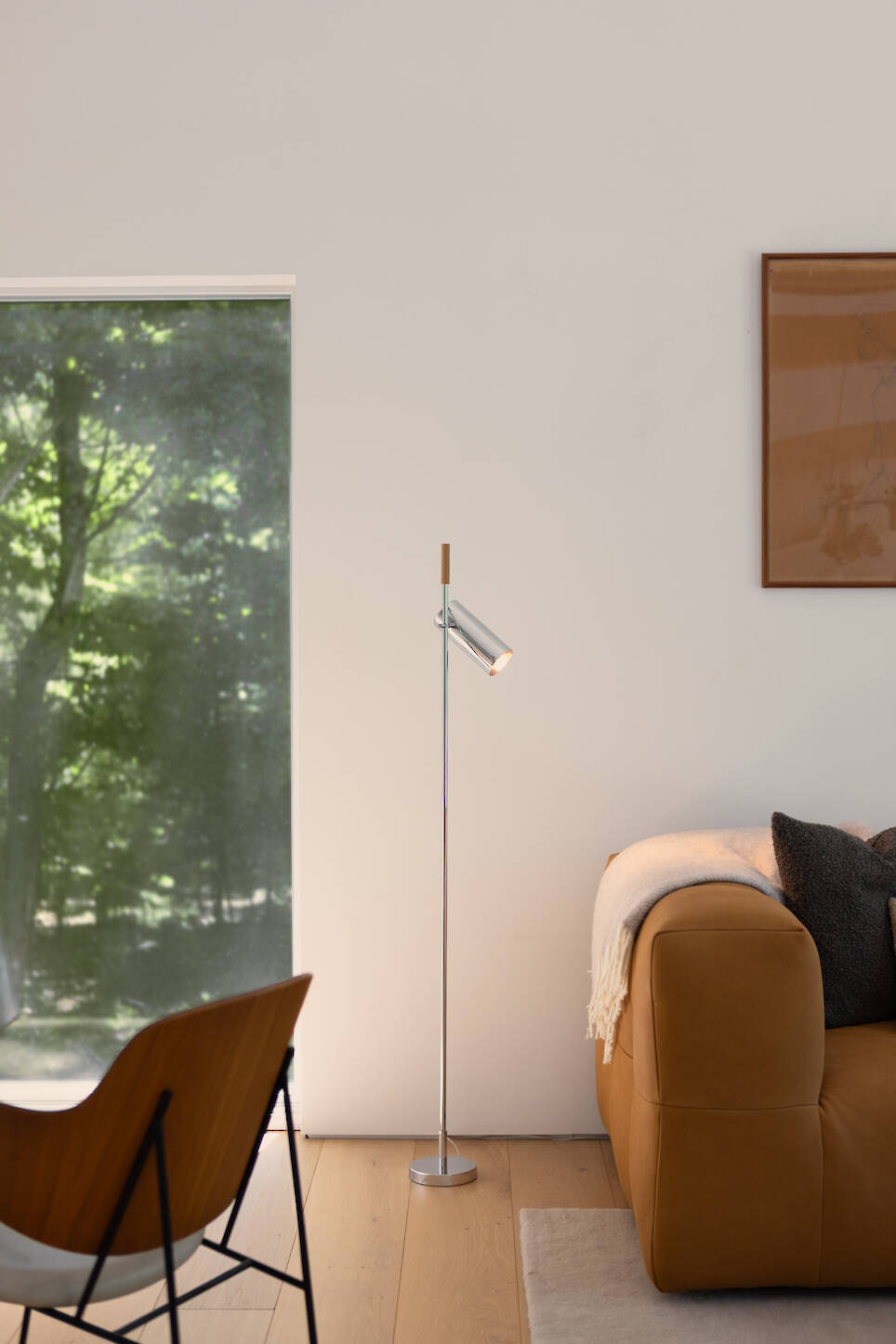 A slender metal floor lamp stands next to a leather sofa in a room with a large window and wood flooring
