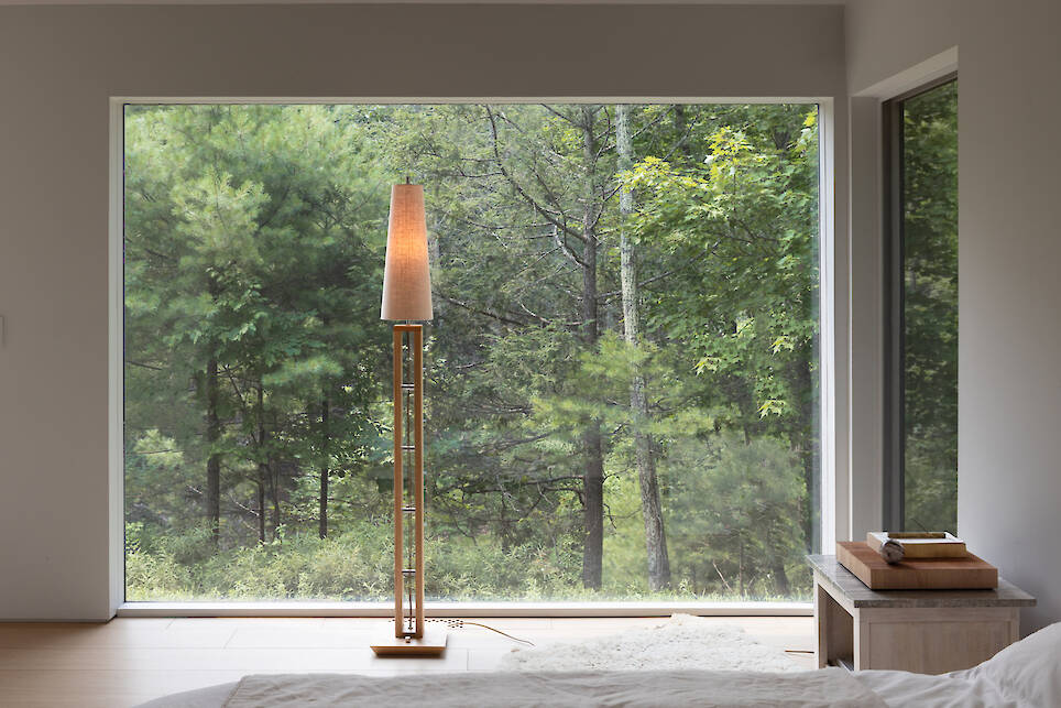 Tall wooden floor lamp with a conical shade in front of a large window overlooking a forest