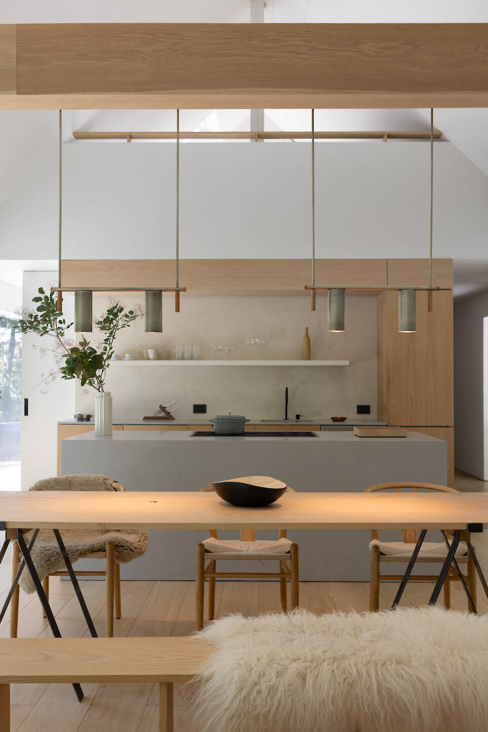 A kitchen island with a cooktop stands in front of a wooden dining table and bench, both topped with sheepskin-style covers, beneath two pendant lights