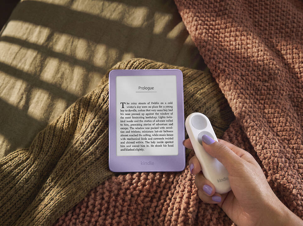 A hand holds a remote next to a purple Kindle redesign e-reader displaying a page of text, resting on folded blankets