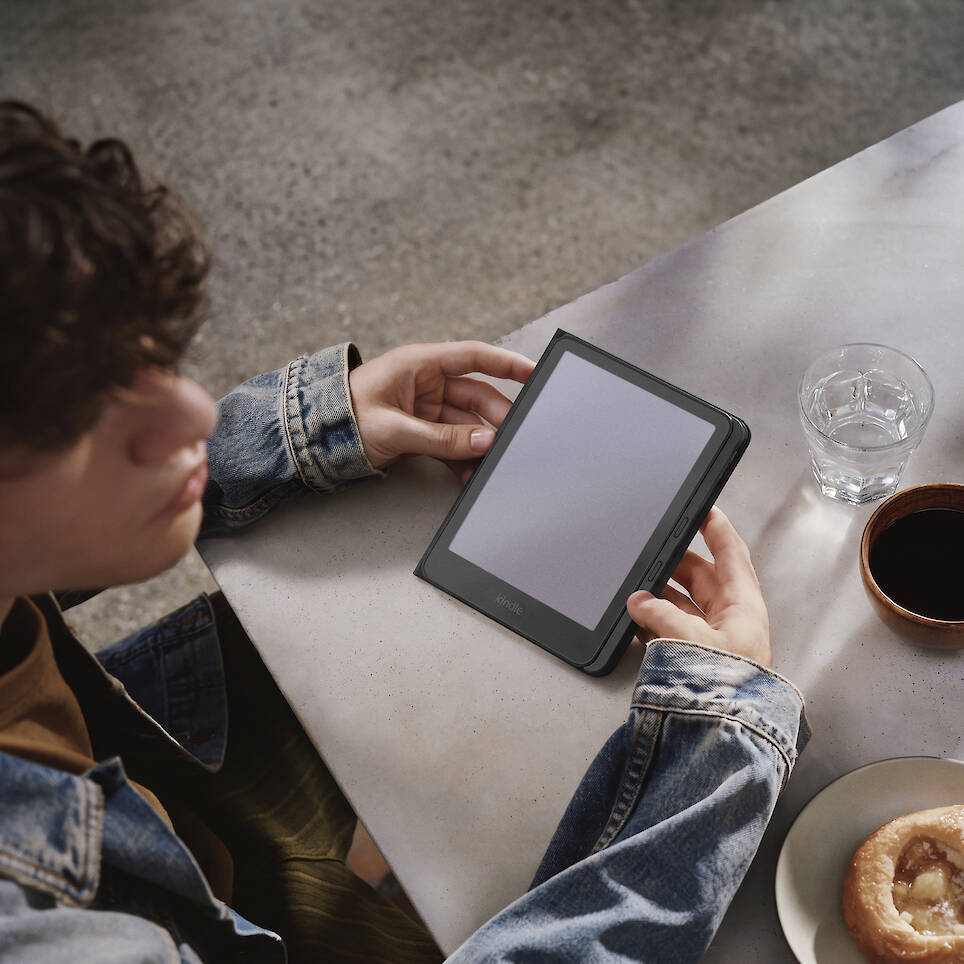 A person in a denim jacket holding an e-reader with a new Kindle redesign at a table with a glass of water, coffee, and a pastry