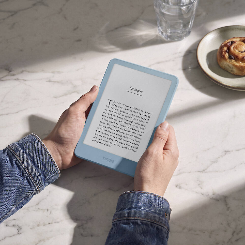 Person holding a light blue Kindle e-reader showcasing a sleek Kindle redesign with the book’s prologue, alongside a pastry and glass of water on a marble surface