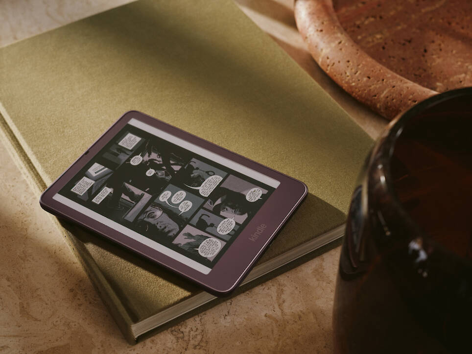 A plum-colored Kindle redesign e-reader displaying a comic book page, resting on a closed olive-green book