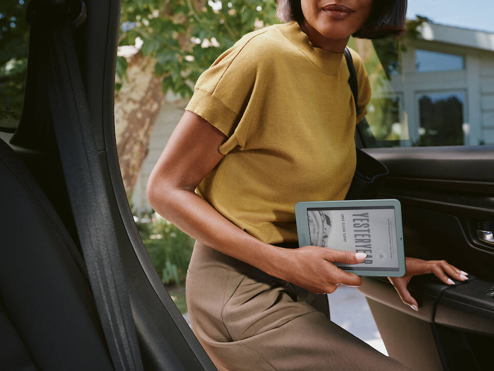 A woman in a mustard shirt exits a car while holding a Kindle redesign e-reader