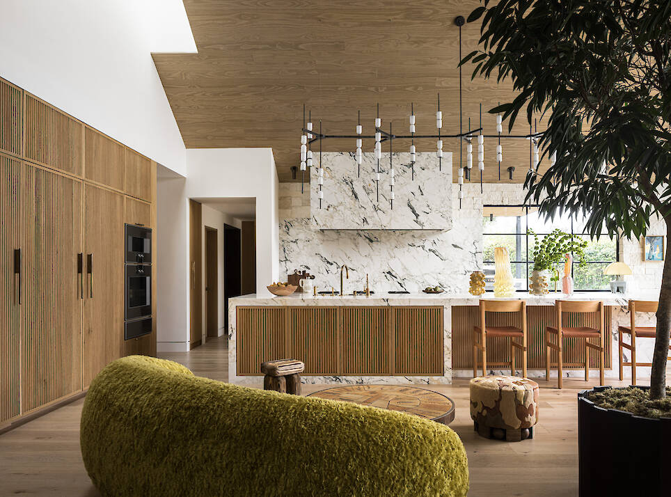 Modern kitchen with a marble island, wood cabinetry, and a black chandelier, viewed from a living area with a large green sofa and a potted tree