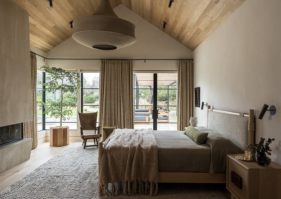 A bedroom with a vaulted wooden ceiling, a bed with a textured headboard, a fireplace, and glass doors opening to a patio