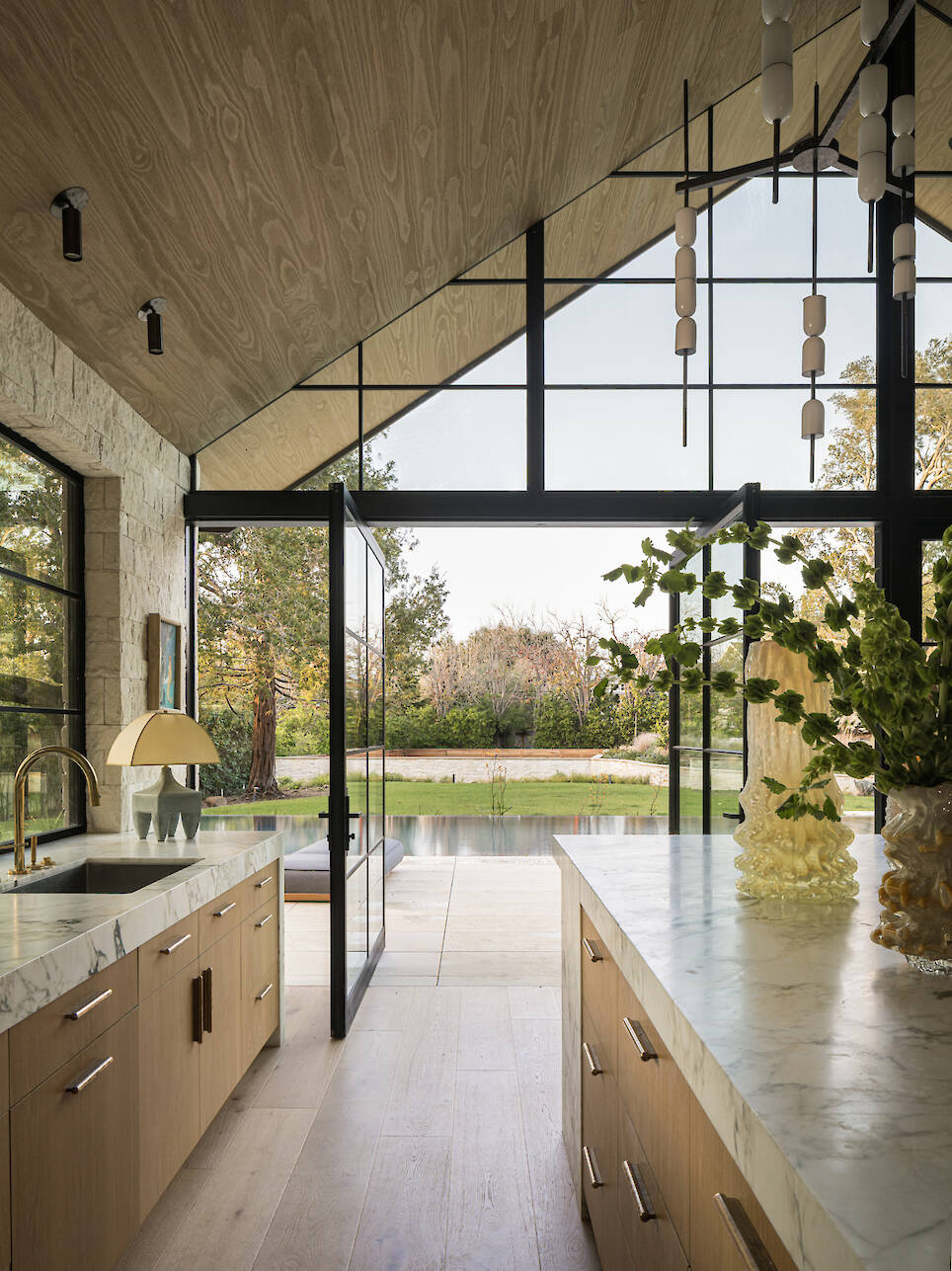 Modern kitchen with light wood cabinets and marble countertops opening onto a patio and swimming pool through large glass doors