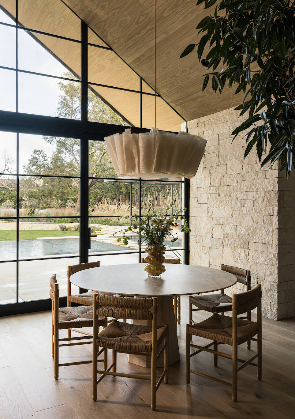 Round dining table with four woven chairs and a flower vase, set in front of floor-to-ceiling windows looking out to a swimming pool