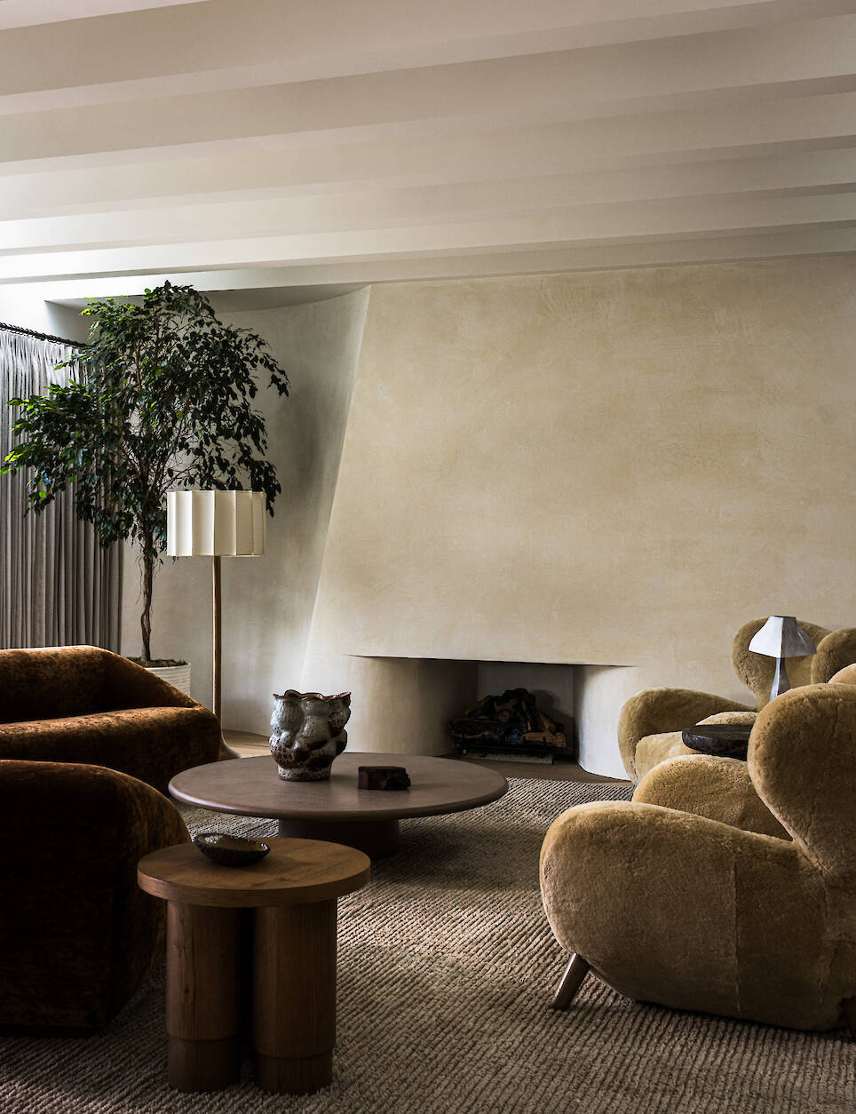 Living room with a large, beige fireplace, curved armchairs, a round coffee table, and a floor lamp next to a large potted plant