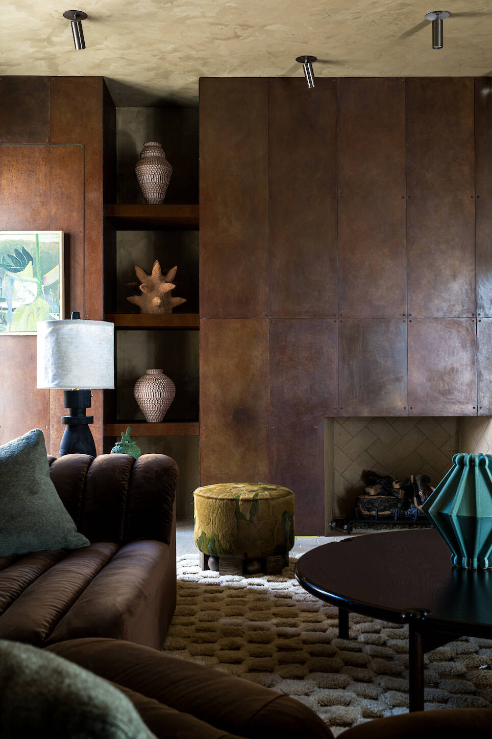 A living room with brown paneled walls, a fireplace, a brown sofa, a patterned ottoman, and decorative vases on shelves
