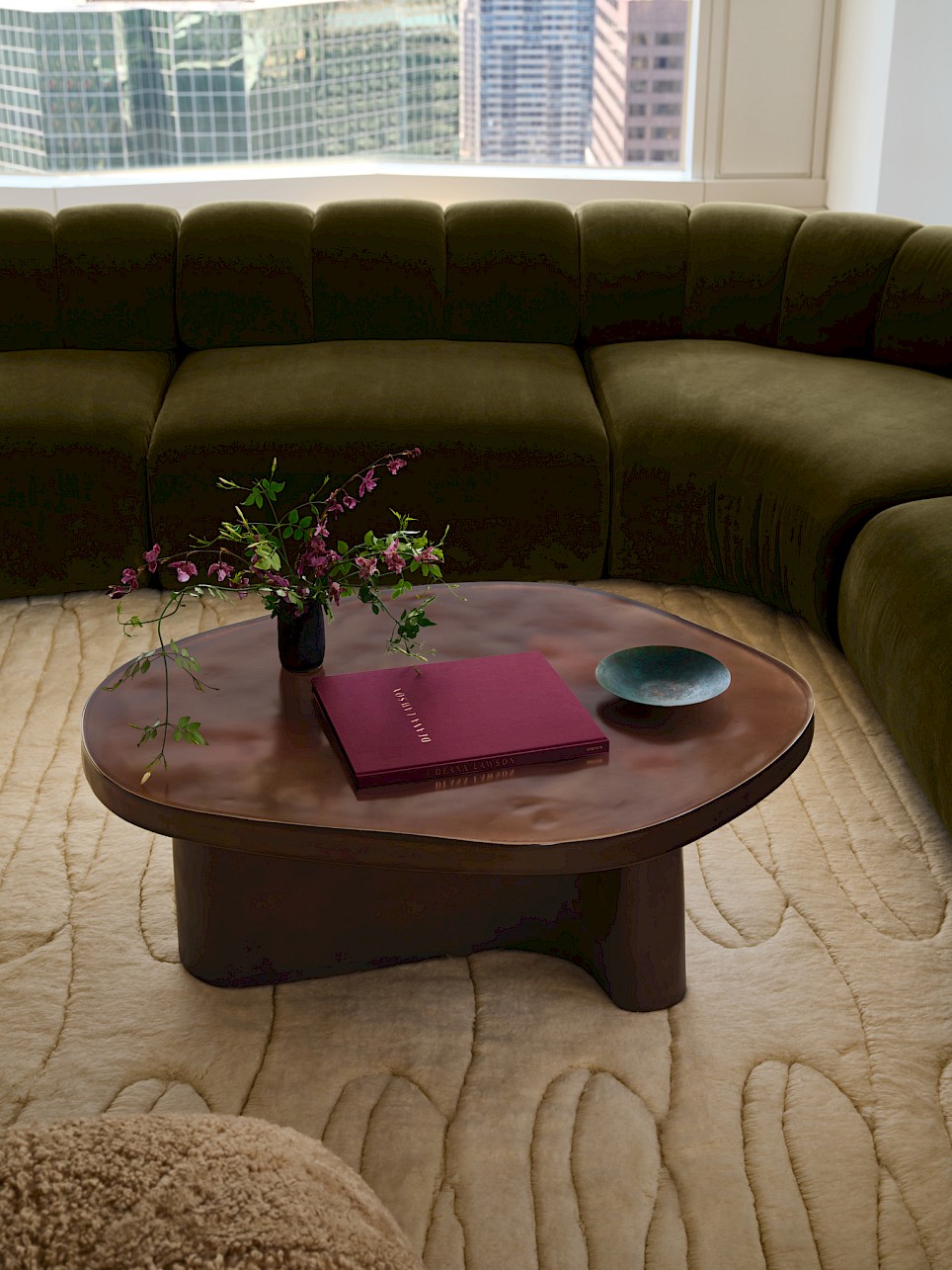 A dark circular Swizz Beatz CB2 coffee table sits in front of a curved olive green sofa, featuring a maroon book, a small vase of flowers, and a shallow bowl