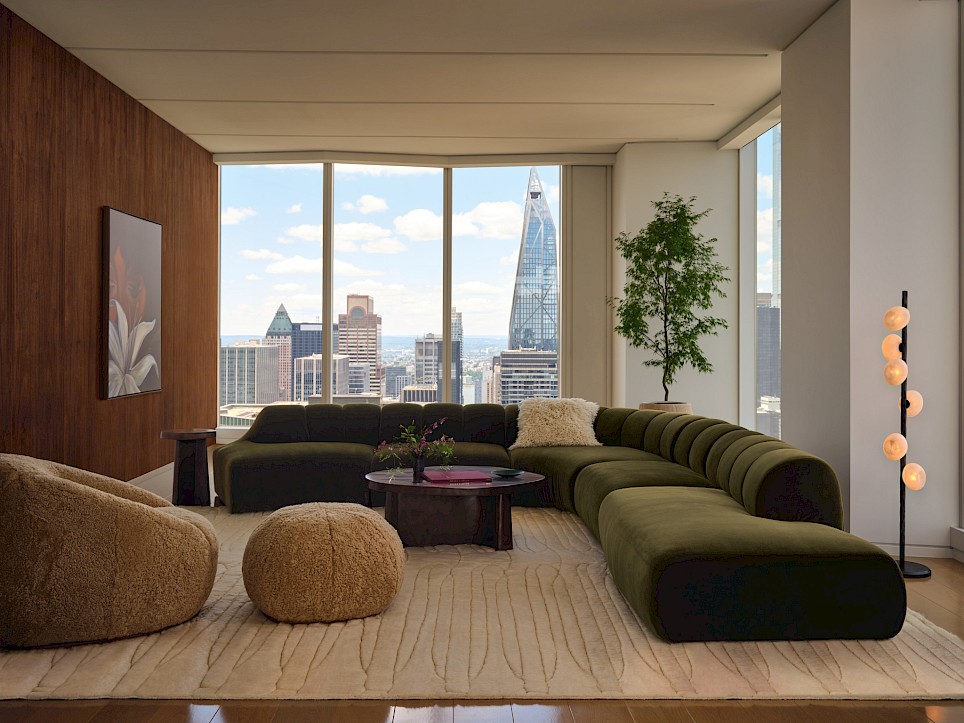 Modern living room featuring a Swizz Beatz CB2 olive green sectional sofa, two textured tan ottomans, and a coffee table, overlooking a city skyline