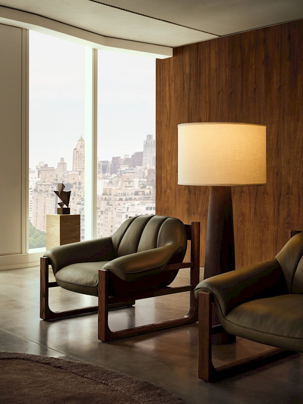 Two olive green Swizz Beatz CB2 leather armchairs and a tall floor lamp in a modern room with a city skyline view through large windows