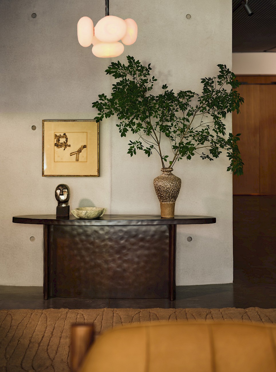 A dark, curved Swizz Beatz CB2 console table holds a metallic head sculpture, a stone bowl, and a tall vase with leafy branches against a concrete wall with a framed print and p