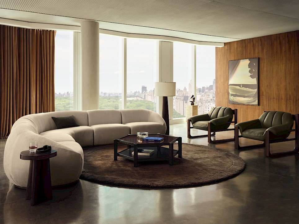 A modern Swizz Beatz CB2 living room with a curved sofa, two armchairs, a coffee table on a circular rug, and floor-to-ceiling windows overlooking a city skyline