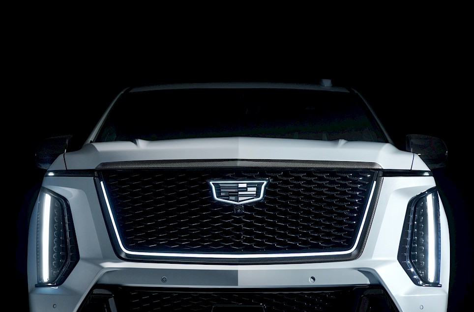 Front view of a white Cadillac Escalade-V 125th Anniversary SUV with an illuminated grille emblem and vertical LED headlights against a black background