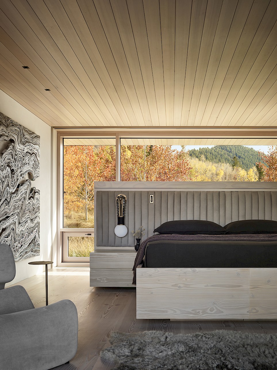 Modern bedroom with a light wood platform bed, upholstered headboard, and large window overlooking an autumn forest