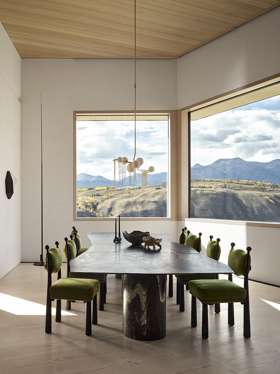 A dining room with a stone-top table and green upholstered chairs, featuring large windows with a view of mountains and a valley