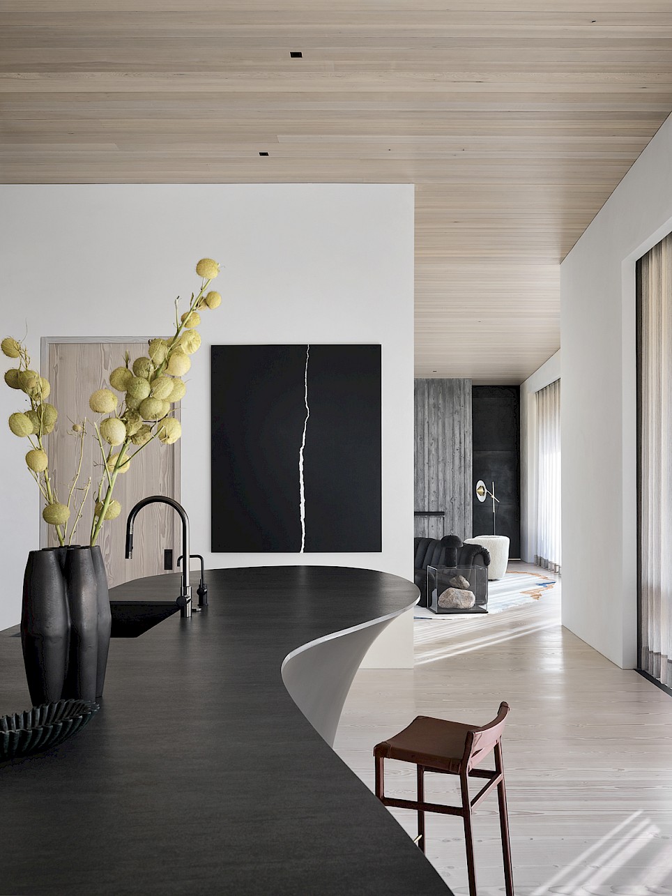 A modern kitchen island with a curved black countertop and a vase of yellow flowers, leading to an open-concept living area with a large abstract black painting