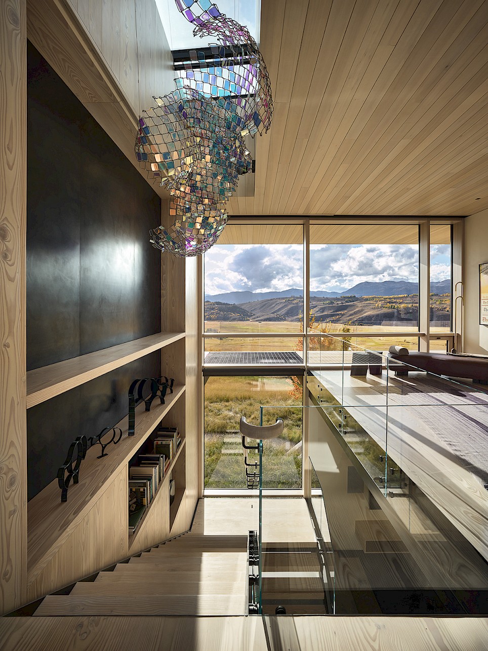 Modern interior with wooden staircase, built-in shelving, a large iridescent sculpture hanging from the ceiling, and a floor-to-ceiling window overlooking a mountain landscape