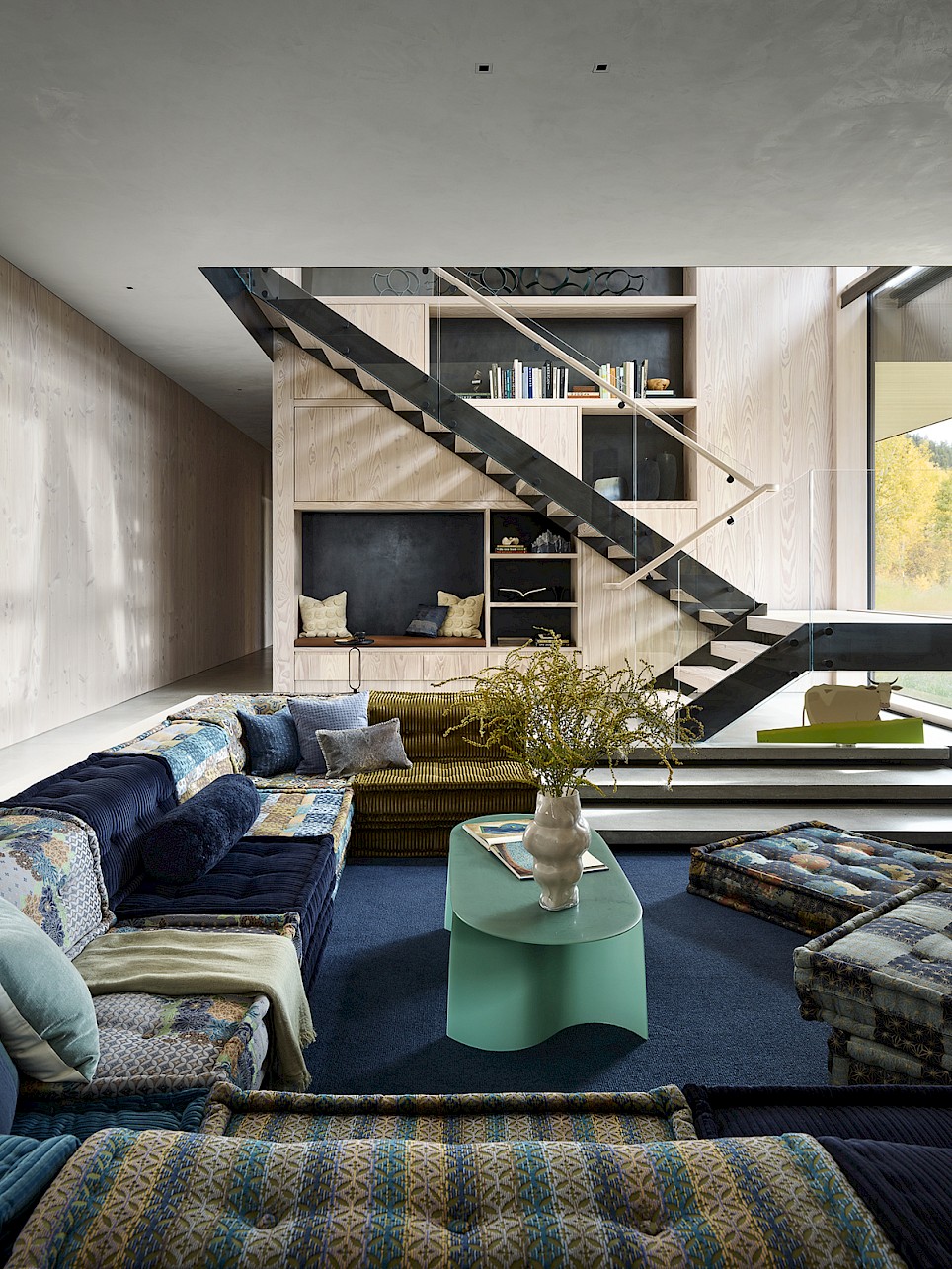 Living room with a large, multi-patterned sectional sofa, a green coffee table, and an open staircase leading to built-in wooden shelving