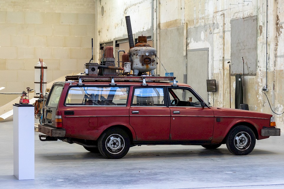 A weathered red station wagon with a custom metal furnace and piping mounted on its roof rack, displayed in an industrial gallery space