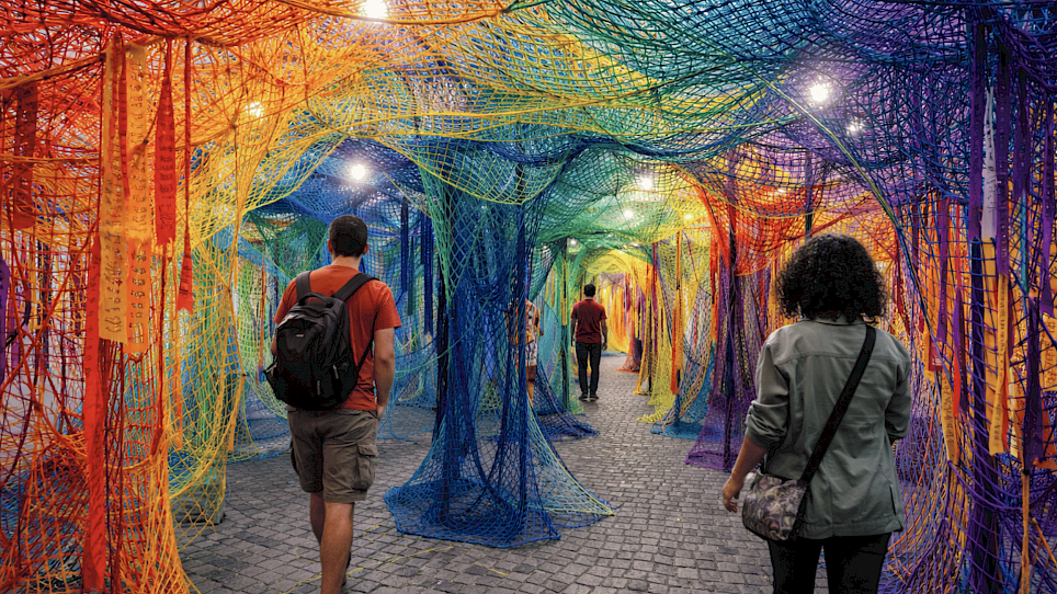 People walk through a path surrounded by a tunnel of multicolored, overlapping mesh nets with dangling ribbons