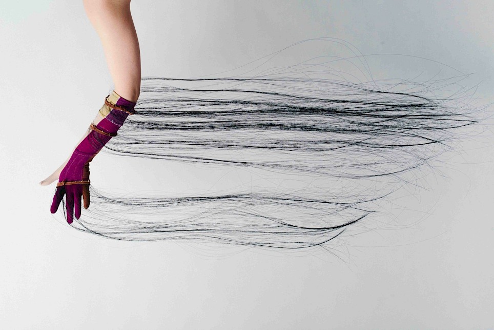 A gloved hand extending horizontally, with long, thin black strands trailing from the fingertips across a white background