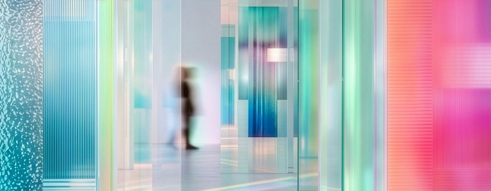 A person walking through a corridor lined with vertical, translucent glass panels in blue, green, and pink gradients
