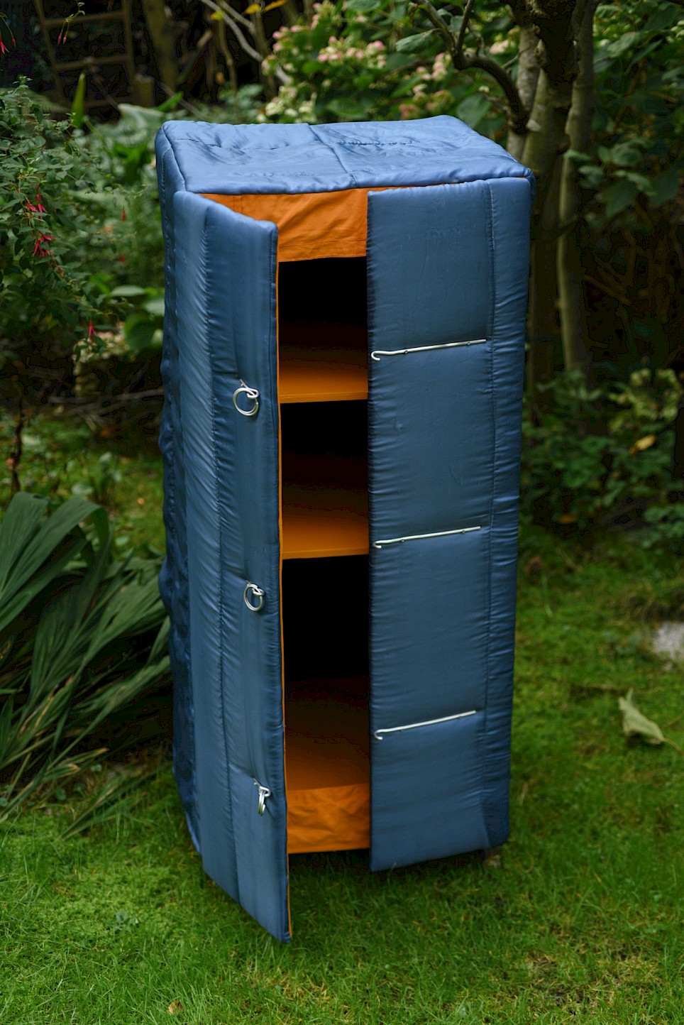 Tall, blue quilted cabinet with an orange interior and three open shelves, standing on grass