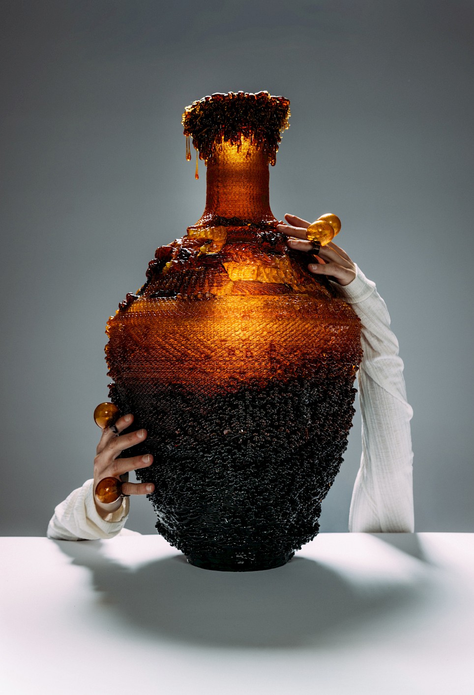 A person in a white long-sleeved shirt holds a large, textured vase that transitions from black at the base to amber at the top, where it appears to be dripping