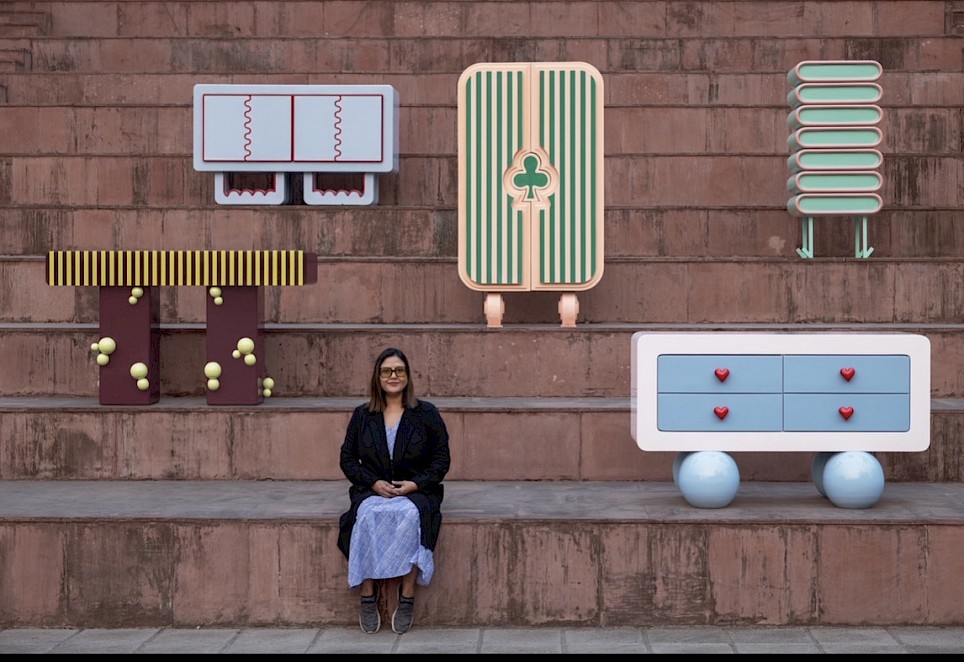A designer sits on stone steps surrounded by five pieces of colorful, stylized furniture