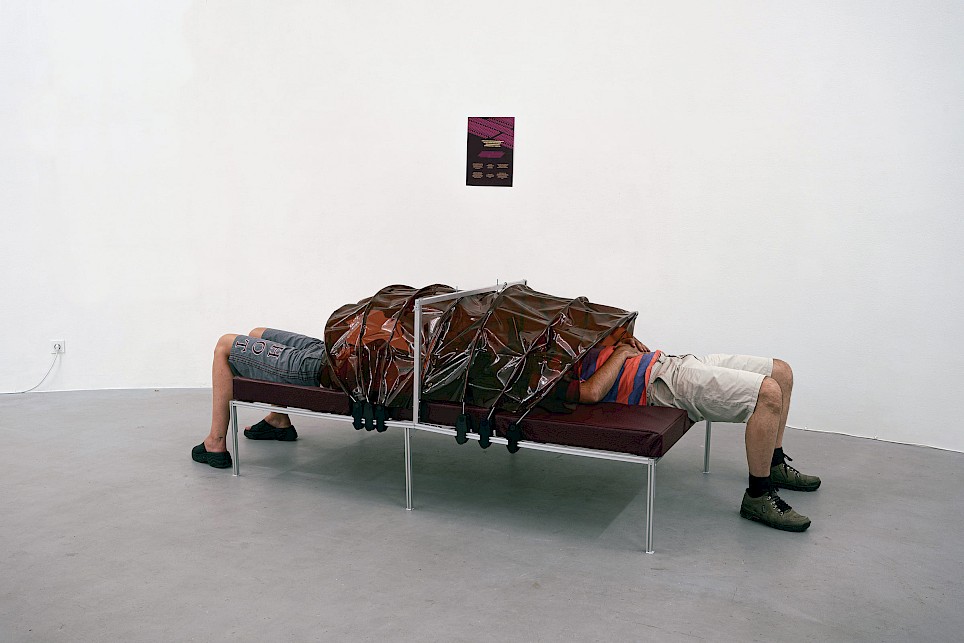 Two people lie at opposite ends of a bench, their upper bodies covered by a brown plastic shroud, with a small poster on the wall behind them