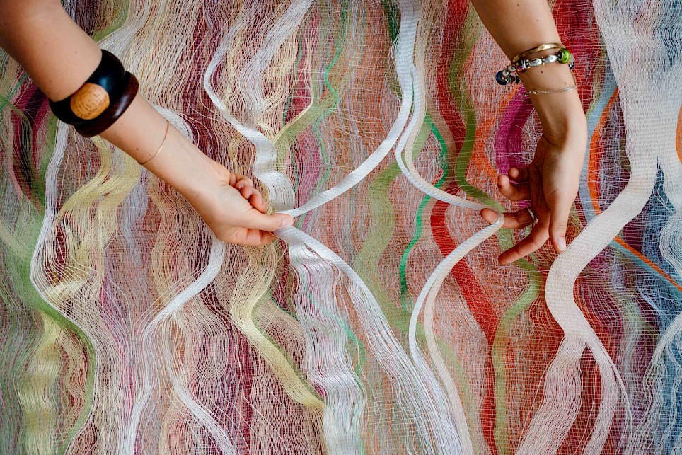 Hands manipulate a sheet of multi-colored, wavy fiber art made of thin, sheer mesh strips