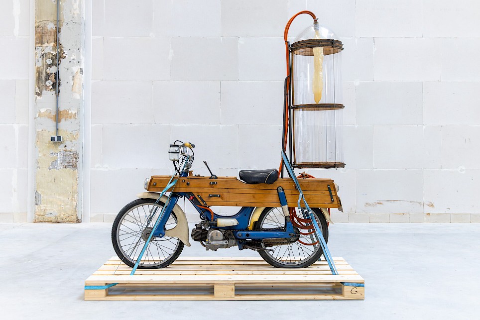 A vintage moped with wood-paneled sides and a tall, cylindrical glass container mounted on the back, standing on a wooden shipping pallet